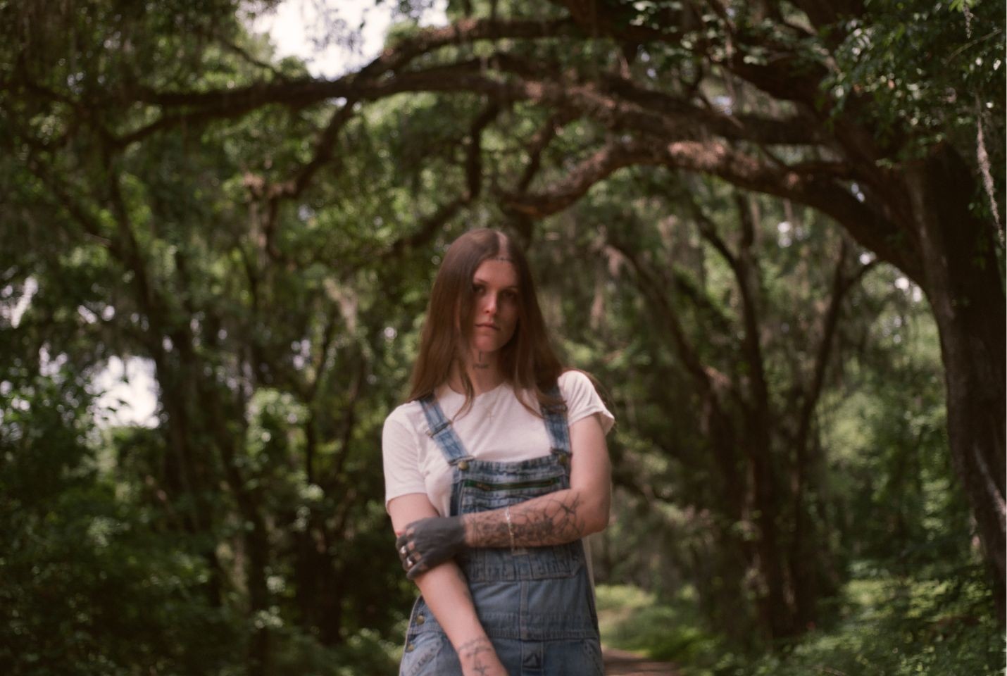 A person with long brown hair wearing a white tshirt and blue denim overalls in a woods